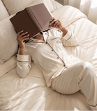 A person lying on a bed reading a brown book, wearing cream-colored pajamas with dark piping and a small embroidered logo on the chest.
